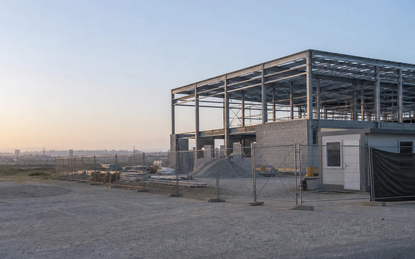 Fenced construction site with a partially completed industrial building and temporary office.